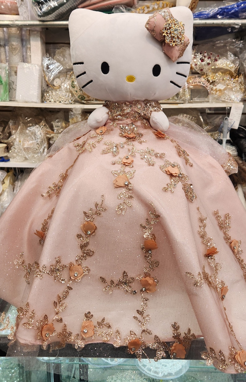 May include: A plush Hello Kitty doll wearing a pink dress with gold glitter and floral embellishments.