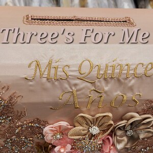 May include: A pink and gold decorative box with the text "Three's For Me" and "Mis Quince A&ntilde;os" embroidered on the front. The box is decorated with gold glitter and lace.