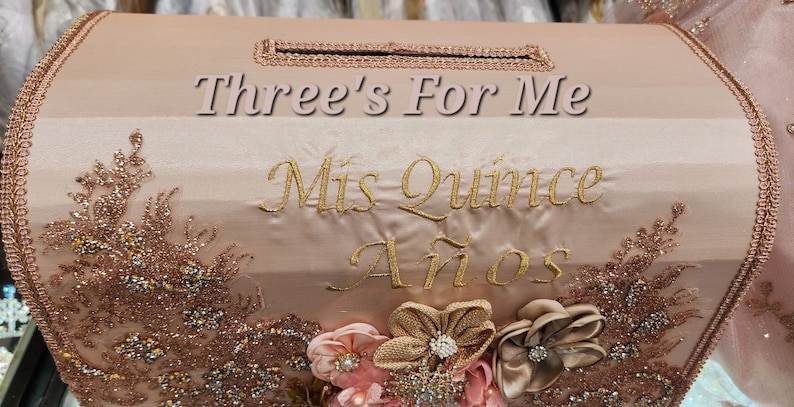 May include: A pink and gold decorative box with the text "Three's For Me" and "Mis Quince A&ntilde;os" embroidered on the front. The box is decorated with gold glitter and lace.