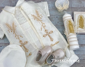 May include: A white christening gown with gold embroidery of wheat stalks, crosses, and the words "Mi Bautizo". The gown is laid out with a white hat, shoes, and a candle with gold embroidery.