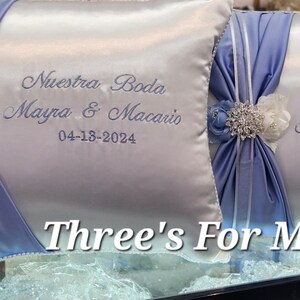 May include: Two white satin pillow covers with blue satin ribbon bows and a rhinestone and flower embellishment. The pillows are embroidered with the text "Nuestra Boda Mayra & Macario 04-18-2024" and "Three's For Me".