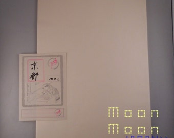 May include: A sheet of off-white paper with the text "MOON MOON JAPAN> PAPER" printed in yellow with a black border. The paper is sitting on a gray surface. A small card with Japanese characters and a drawing of a person is in the lower left corner of the image.