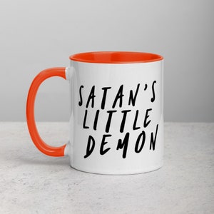 May include: White ceramic mug with an orange interior and handle. The mug features the text "SATAN'S LITTLE DEMON" in black, handwritten-style lettering. The mug is designed for hot beverages.
