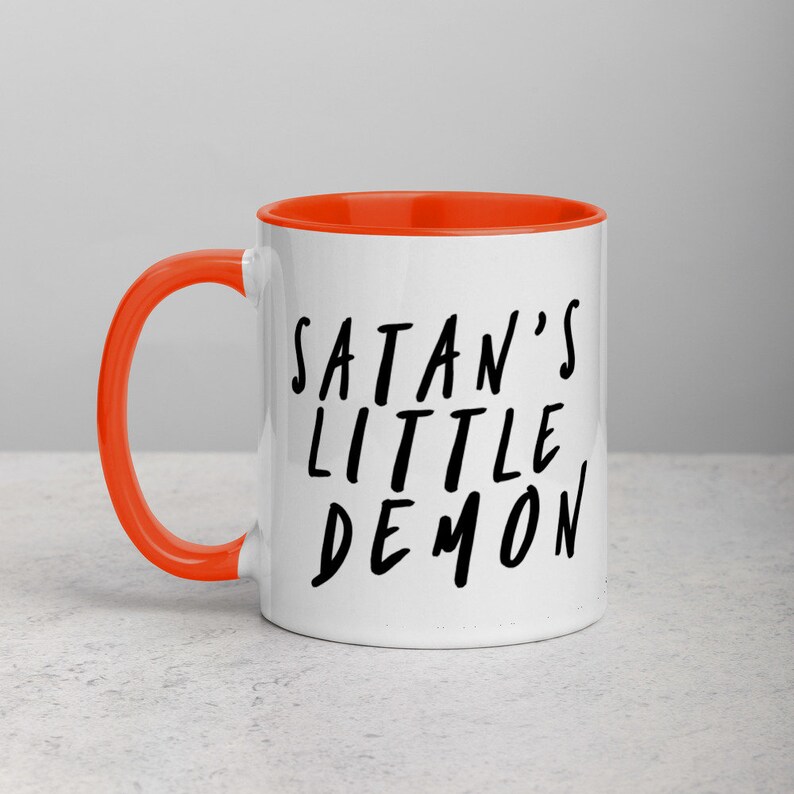 May include: White ceramic mug with an orange interior and handle. The mug features the text "SATAN'S LITTLE DEMON" in black, handwritten-style lettering. The mug is designed for hot beverages.