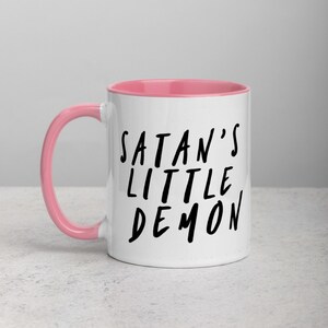 May include: White ceramic mug with a pink interior and handle. The mug features the black text "SATAN'S LITTLE DEMON" in a handwritten style. The mug is designed for hot beverages.