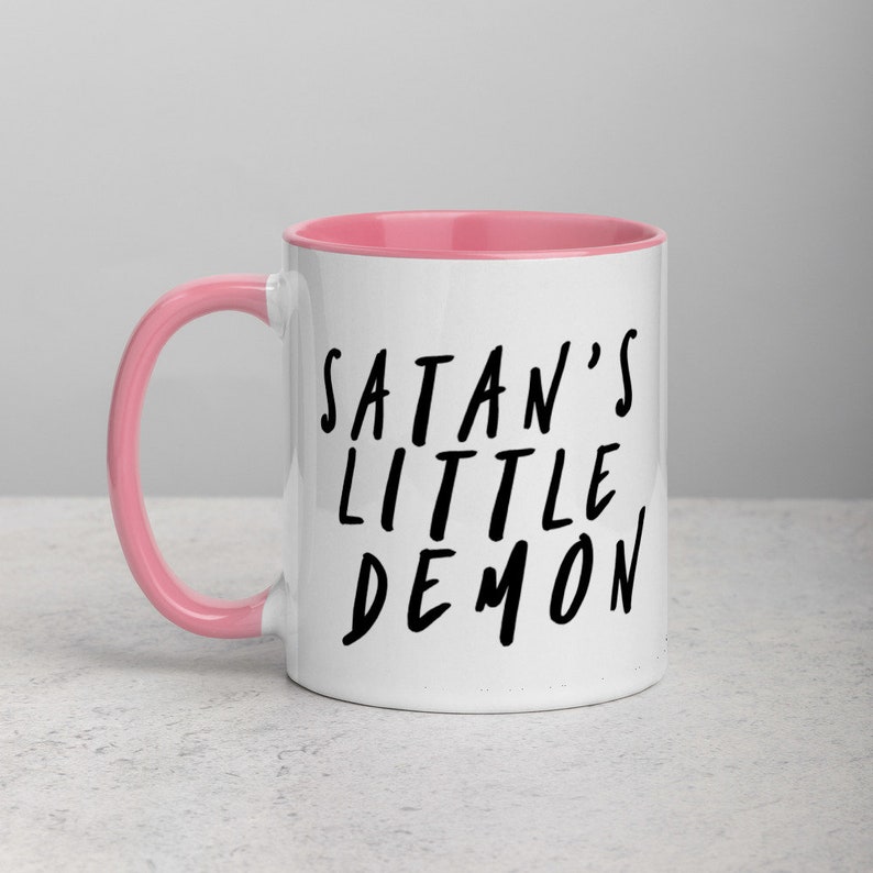 May include: White ceramic mug with a pink interior and handle. The mug features the black text "SATAN'S LITTLE DEMON" in a handwritten style. The mug is designed for hot beverages.