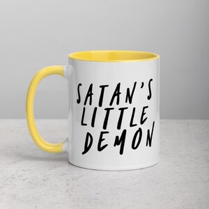 May include: White ceramic mug with a yellow interior and handle. The mug features the text "SATAN'S LITTLE DEMON" in black, handwritten-style lettering. The mug is on a light gray surface.