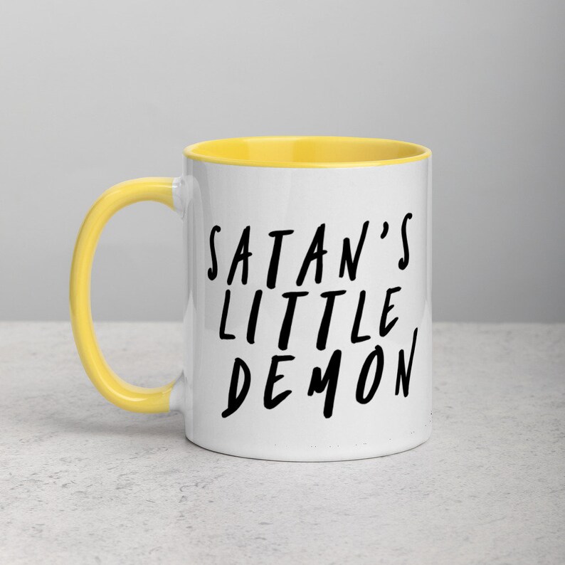 May include: White ceramic mug with a yellow interior and handle. The mug features the text "SATAN'S LITTLE DEMON" in black, handwritten-style lettering. The mug is on a light gray surface.