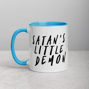May include: White ceramic mug with a light blue interior and handle. The mug features the text "SATAN'S LITTLE DEMON" in black, handwritten-style lettering. The mug is designed for hot beverages.