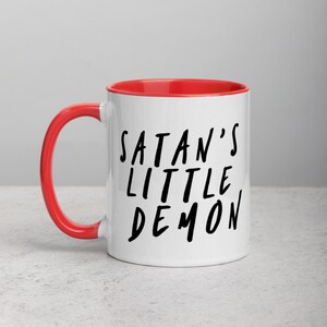 May include: White ceramic mug with a red interior and handle. The mug features the text "SATAN'S LITTLE DEMON" in black, handwritten-style lettering. The mug is set against a neutral background.