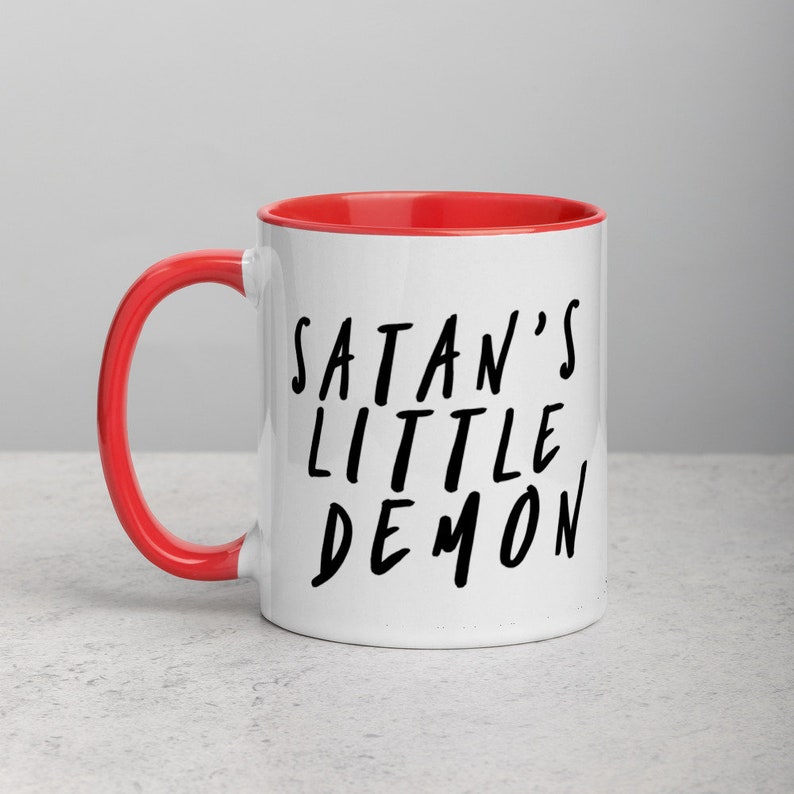 May include: White ceramic mug with a red interior and handle. The mug features the text "SATAN'S LITTLE DEMON" in black, handwritten-style lettering. The mug is set against a neutral background.