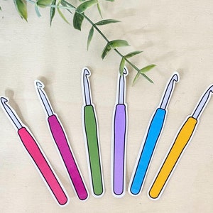 May include: Seven colorful crochet hooks, each with a different color handle: pink, magenta, green, purple, blue, and yellow. The hooks are arranged in a row on a light wood surface.