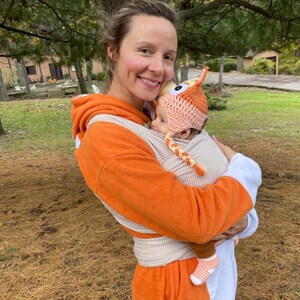 May include: A woman wearing an orange and white striped baby carrier with a baby wearing a crocheted fox hat. The woman is smiling and the baby is looking at the camera.