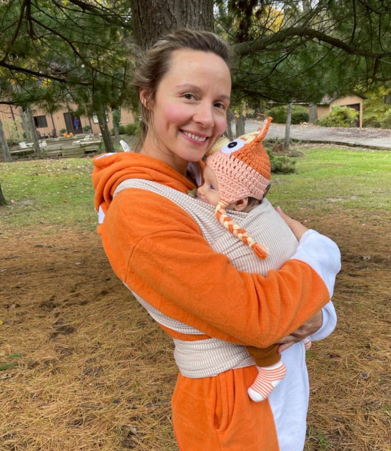 May include: A woman wearing an orange and white striped baby carrier with a baby wearing a crocheted fox hat. The woman is smiling and the baby is looking at the camera.