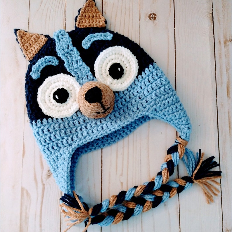 May include: Blue and brown crocheted hat with a dog face design. The hat has a long braided tail and ear flaps.