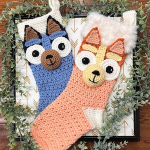 May include: Two handmade crochet Christmas stockings. One is blue with brown accents, the other is peach with orange accents. Both have white fur trim and are decorated with cartoon dog faces.
