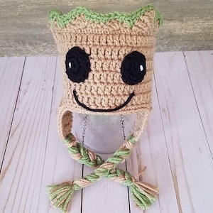 May include: A crocheted hat in the shape of a character with a smiling face, black eyes, and a green leafy top. The hat has ear flaps and braided ties with tassels. The hat is beige, green, and black.