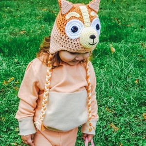 May include: A child wearing a crocheted fox hat with orange, white, and yellow yarn. The hat has large white eyes and a brown nose. The child is wearing a peach colored sweatshirt and pants.