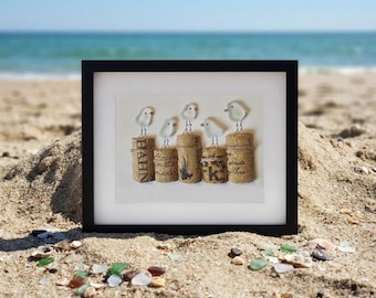 May include: A framed wall art piece featuring five white sea glass birds perched on five upright wine corks. The corks have printed text and graphics on their sides. The birds have thin black ink legs and eyes drawn onto the white glass pieces.