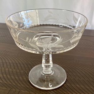 May include: A clear, stemmed glass dessert bowl with etched fern leaf designs. The bowl has a wide, shallow top and a round base. The etched details add a delicate touch to the glassware, suitable for serving desserts or displaying small items.