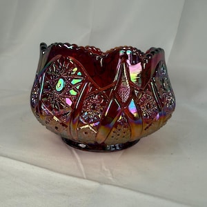 May include: A ruby red, iridescent glass bowl with a scalloped rim. The bowl features a detailed pattern of stars, diamonds, and floral designs. The glass has a shimmering, oil-slick effect, with hints of green, blue, and gold. The bowl is likely vintage or antique.
