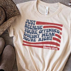 May include: A natural-colored t-shirt with the text "JUST BECAUSE YOU'RE OFFENDED DOESN'T MEAN YOU'RE RIGHT" in blue and red, resembling a flag. The text "Charlie Kirk" is below the flag design.