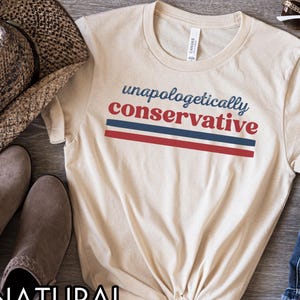 May include: A cream-colored t-shirt with the words "unapologetically conservative" in blue and red. The text is above two horizontal stripes in red and blue. The shirt is folded at the bottom. A hat and boots are visible in the background.