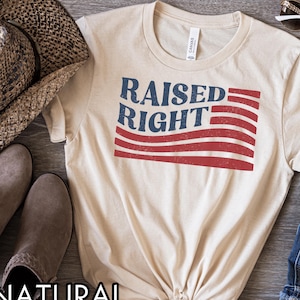 May include: A beige t-shirt with the words "RAISED RIGHT" in blue and red, with a flag design. The shirt is made of a soft material and is likely a unisex style. The shirt is folded at the bottom.