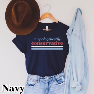 May include: Navy blue t-shirt with the text "unapologetically conservative" in blue and red. Below the text are three horizontal stripes in blue, white, and blue. A brown hat hangs above the shirt, and a light blue denim shirt is draped on the right.