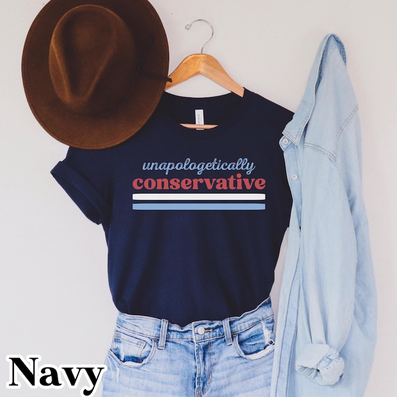 May include: Navy blue t-shirt with the text "unapologetically conservative" in blue and red. Below the text are three horizontal stripes in blue, white, and blue. A brown hat hangs above the shirt, and a light blue denim shirt is draped on the right.