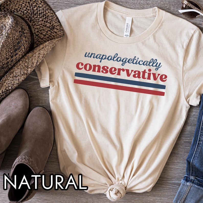 May include: A cream-colored t-shirt with the words "unapologetically conservative" in blue and red. The text is above two horizontal stripes in red and blue. The shirt is folded at the bottom. A hat and boots are visible in the background.