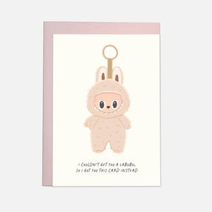 Könnte beinhalten: Eine Grußkarte mit einem hellrosa Umschlag. Die Karte zeigt eine Cartoonfigur in einem Hasenkostüm mit einem Schlüsselanhänger. Der Text auf der Karte lautet: "I COULDN'T GET YOU A LABUBU, SO I GOT YOU THIS CARD INSTEAD."