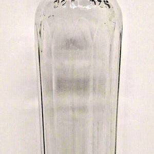 May include: A clear glass bottle with a tall, slender neck and a rounded rim. The body features vertical fluted panels and is embossed with the text 32 FL OZS near the shoulder. The bottle has a circular base and a transparent, smooth finish.