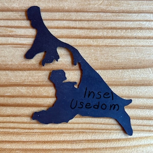 Puede incluir: Una silueta negra de la isla de Usedom, Alemania, con el texto "Insel Usedom" escrito en ella. La silueta est&aacute; sobre una superficie de madera.