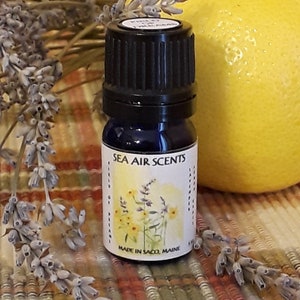 May include: A small blue glass bottle with a black cap and a white label that reads "SEA AIR SCENTS" and "MADE IN SACO, MAINE". The bottle is sitting on a plaid surface with lavender flowers.