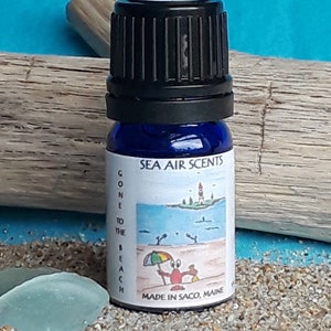 May include: A small blue glass bottle with a black lid. The label reads "Sea Air Scents" and "Gone to the Beach". There is a cartoon image of a lighthouse, a beach, and a crab holding an umbrella.