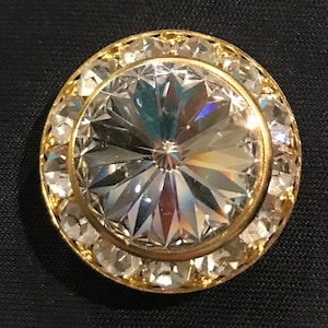 May include: A large, clear crystal button with a gold setting and a surrounding halo of smaller crystals.