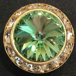 May include: A round, decorative button with a central, faceted, light green gemstone. The gemstone is surrounded by a gold-colored metal band, which is encircled by a ring of clear, sparkling crystals. The button is set against a dark background.