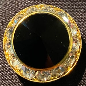 May include: A round, black onyx cabochon set in a gold tone metal frame with a border of clear rhinestones.