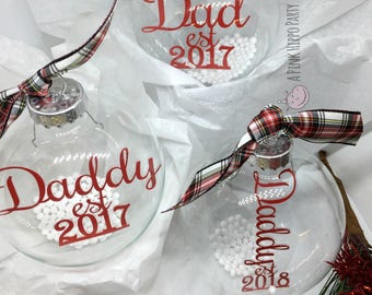 May include: Three clear glass Christmas ornaments with red ribbon and white beads inside. The ornaments are decorated with red vinyl lettering that reads "Dad est 2017", "Daddy est 2017", and "Daddy est 2018".