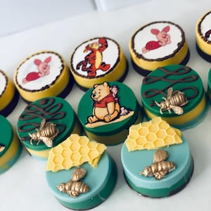 May include: A collection of decorated round treats. Some feature Winnie the Pooh characters like Piglet and Tigger, while others have bee and honeycomb designs. The treats have yellow, green, and blue layers with gold accents.