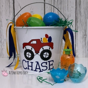 May include: A white metal bucket with a monster truck design and the name "CHASE" printed on it. The bucket is filled with plastic Easter eggs and green grass. There are also two blue plastic Easter egg holders and a clear plastic container with a green lid.