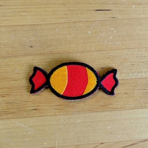 May include: A red and yellow candy shaped patch with black outlines. The patch is on a wooden surface.