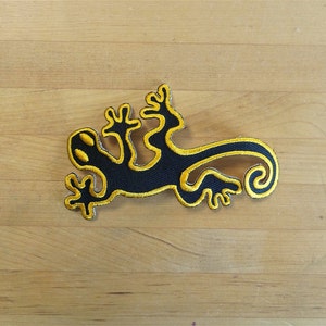 May include: Black and yellow embroidered gecko patch. The gecko is outlined in yellow and has a curled tail.