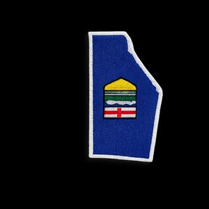 Könnte beinhalten: Ein blaues, gesticktes Abzeichen in Form einer kanadischen Provinz. Der Aufnäher hat einen weißen Rand und ein zentrales Emblem mit gelben, grünen, weißen und roten Elementen. Der Aufnäher ist vor einem schwarzen Hintergrund platziert.
