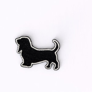 Peut inclure: Écusson noir en forme de chien avec un contour brodé blanc. L'écusson présente une silhouette détaillée d'un chien, avec des pattes, une queue et une tête visibles. L'écusson est probablement destiné aux vêtements ou aux accessoires.
