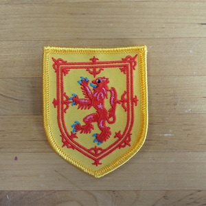 Op de afbeelding: Een gele geborduurde patch met een rode leeuw die op een gele achtergrond staat. De leeuw is een heraldisch symbool van Schotland.