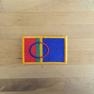 Op de afbeelding: Een geborduurde patch in geel, rood, groen en blauw met een cirkel in het midden. De cirkel is blauw met een gele rand. De patch is omgeven door een gele rand.