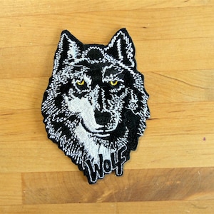 Op de afbeelding: Zwarte en witte geborduurde patch van een wolvenkop met gele ogen. Het woord "Wolf" is onder het gezicht geborduurd.