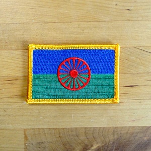 May include: Embroidered patch featuring the Romani flag. The flag design includes a blue top, green bottom, and a red chakra in the center. The patch has a yellow border and is rectangular in shape.